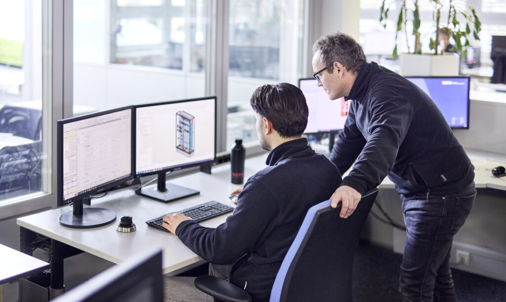 Zwei Männer, einer sitzend am Schreibtisch, der andere dahinter stehend beraten sich gemeinsam bei der Konstruktion am Computer.