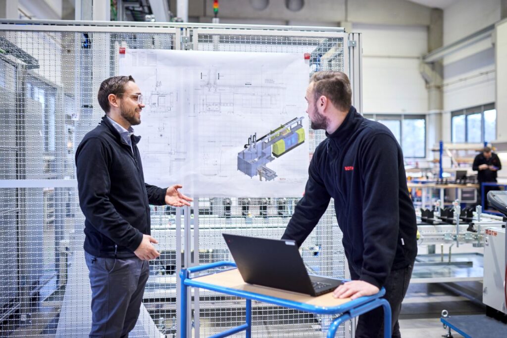 Zwei Männer besprechen sich vor einer großen technischen Zeichnung mit dem Laptop