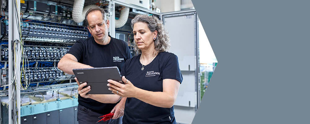 Ein Mann und eine Frau mit einem Tablet in der Hand beraten sich vor einem großen Schaltschrank.