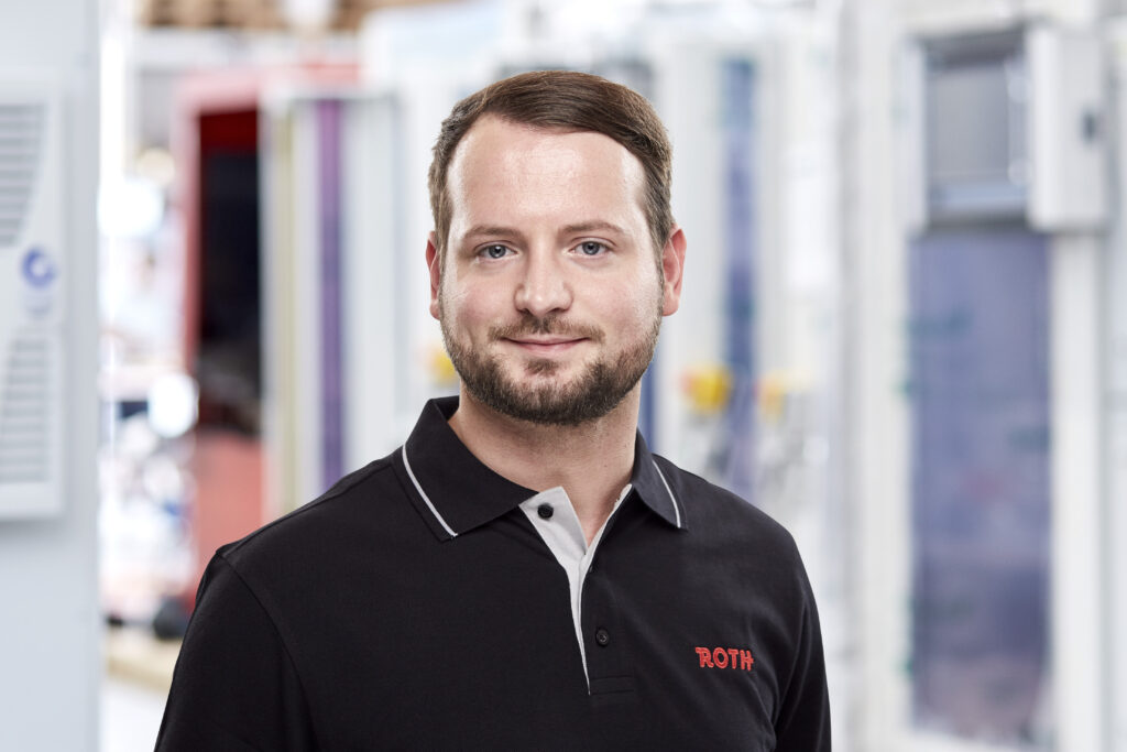 Ein Mann steht im Vordergrund, während im Hintergrund leicht verschwommene Schaltschrankanlagen zu sehen sind