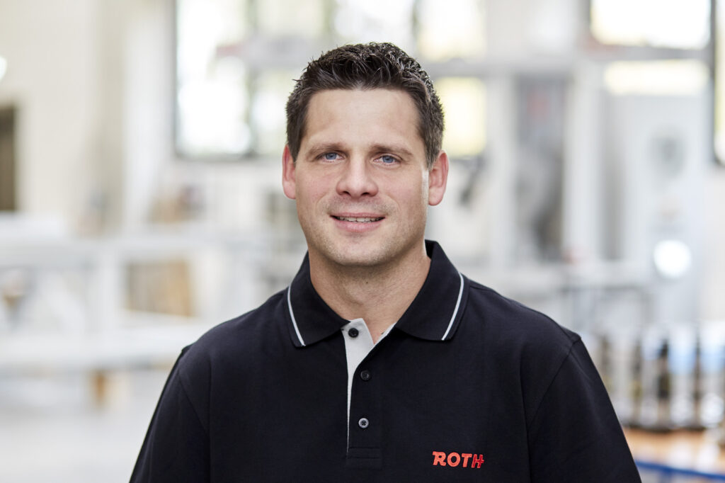 Ein Mann steht im Vordergrund, während im Hintergrund leicht verschwommene Schaltschrankanlagen zu sehen sind