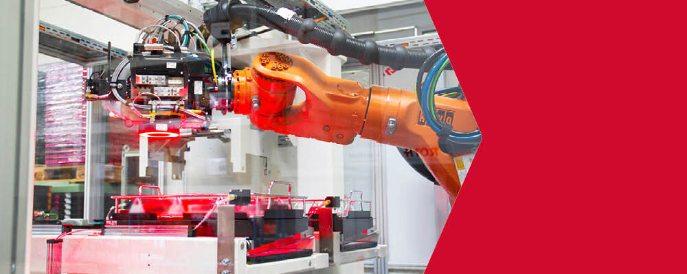 Oranger Industrieroboter hinter einer Schutzglasscheibe, der Werkstücke in einer automatisierten Produktionsanlage umsetzt.